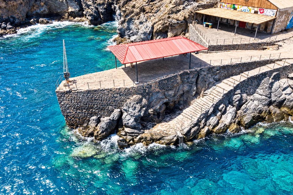 A stone jetty with a red roof beside the turquoise sea, seen from above